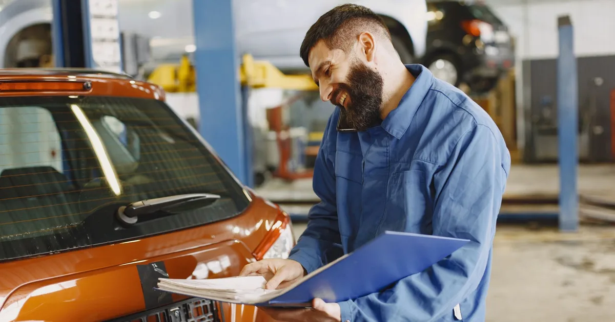 MOT certificate explained: mechanic in a garage checking a car's MOT record on a clipboard beside a lifted vehicle